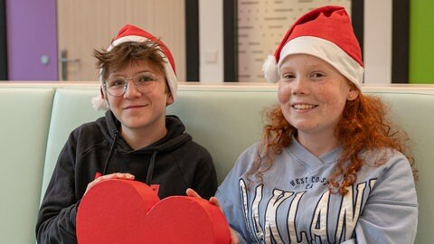 Ein Junge mit Brille und ein Mädchen mit Sommersprossen tragen Weihnachtsmützen und halten zusammen ein Herz.